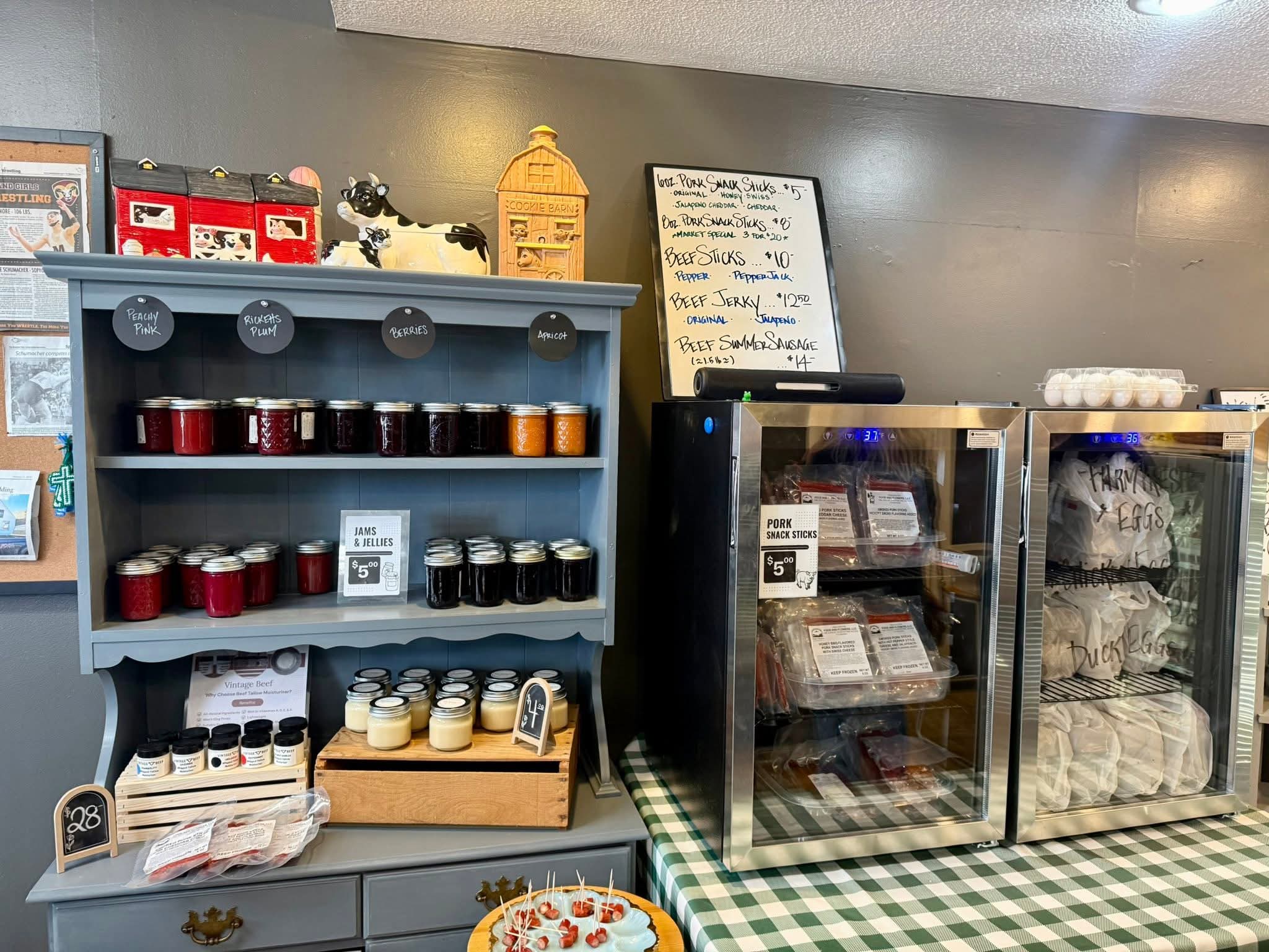 A rustic shop counter featuring a blue-gray wooden shelf stocked with jars of preserves and spreads, alongside two stainless steel glass-door refrigerators, a white handwritten menu board, and a green-and-white checkered tablecloth.